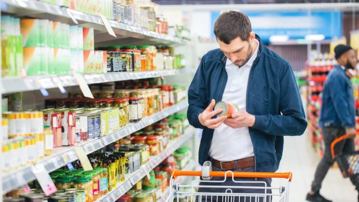 Au supermarché: Bel homme utilise un smartphone et examine la valeur nutritionnelle des conserves. Il est debout avec le panier dans la section des conserves.